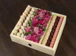 On the left, savor the decadence of Chocolate Covered Strawberries and Chocolate Covered Bananas, a classic and irresistible combination. In the center, the box showcases beautiful flowers, adding a touch of natural beauty and fragrance. On the right, discover a variety of exquisite macarons, each a burst of flavor and color.